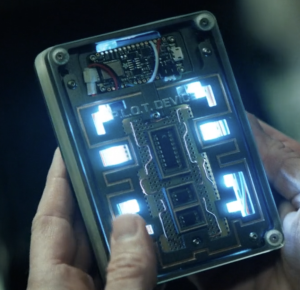 Male hands holding a rectangular clear plastic box with circuit boards, lights and chips in it. It is labeled "P.L.O.T. Device"