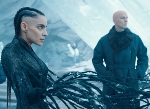 A young Venezuelan woman with black braided hair, a close-fitting plackish outfit and tendrils coming out of her hands in front of a icy white mountain background. A young bald man in a fur coat is standing to the mid-back of the image.